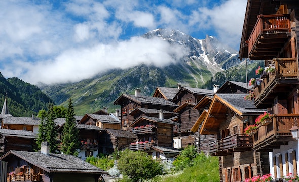 A green mountain with snowy peaks that has wooden cabins at the base.